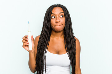Fototapeta premium Young African American woman holding electric toothbrush isolated on blue background confused, feels doubtful and unsure.