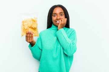 Young African American woman holding potato chips isolated on blue background biting fingernails,...