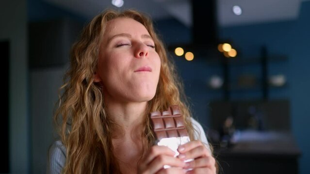Hungry woman bites into a bar of chocolate and enjoys snack. Curly blonde girl overeats unhealthy sweet food. Female stuffs her face into candy at kitchen interior. Healthy diet, overweight concept.