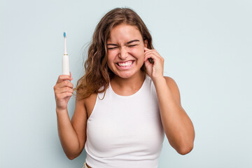 Young caucasian woman holding electric toothbrush isolated blue background covering ears with hands.