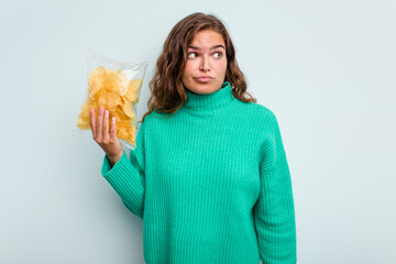 Young caucasian woman holding potato crips isolated on blue background confused, feels doubtful and...