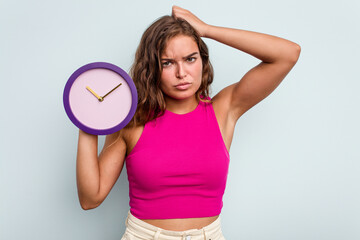 Young caucasian woman holding a clock isolated on blue background being shocked, she has remembered important meeting.