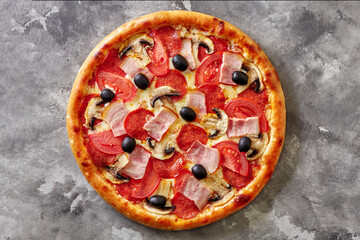 Thin pizza with bacon, salami, mushrooms, tomatoes and black olives on gray stone surface