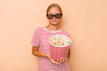 Little caucasian girl eating popcorn isolated on beige background shrugs shoulders and open eyes confused.