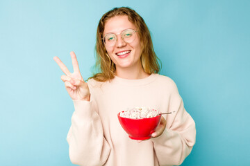 Young caucasian woman holding a bowl of cereals isolated on blue background joyful and carefree showing a peace symbol with fingers.