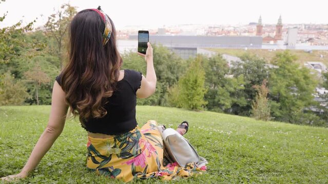 A young beautiful Caucasian woman takes pictures of a cityscape as she sits on a grassy hill