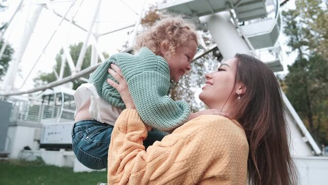Loving mother walks with her cute daughter in the park in sweaters. She holds her daughter in her arms, smiles. Ferris wheel in the background. loving family
