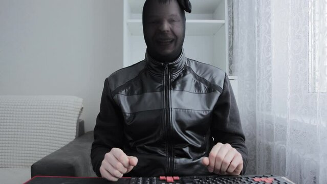 A hacker and internet scammer in a black stocking on his head smiles at the camera in front of a keyboard and computer. Concept of cyber attack, scams, hacking sites. Theft of personal data