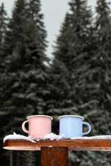 Obraz na płótnie Canvas Two coffee camping mug in winter forest. Couple metal cups with warm drink in pastel pink and blue on background of green fir tree. Blank, mockup, copy space for design, text or promotional content