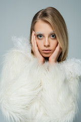 blonde young woman in stylish faux fur jacket with white feathers looking at camera isolated on grey.