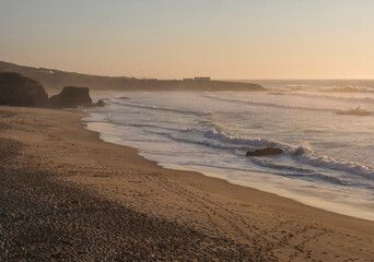 View of empty sand beach Praia Grande de Almograve with ocean waves in golden hour light, clear blue sky. Rota Vicentina coast, Almograve, Portugal