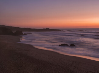 Mysterious long exposure view of sand beach Praia Grande de Almograve with blurred ocean waves in pink orange and purple light. Blue hour after sunset at Rota Vicentina coast, Almograve, Portugal