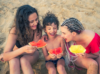 family, leisure and people concept - happy mother and two children having picnic on summer beach and eating red and yellow watermelon