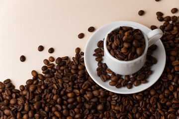 Espresso cup filled with coffee beans surrounded by a large number of coffee beans on a light beige background close-up.