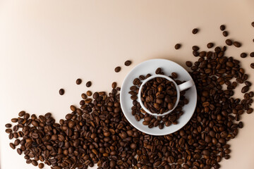 Espresso cup filled with coffee beans surrounded by a large number of coffee beans on a light beige background close-up.