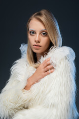 young blonde woman with winter makeup looking at camera and posing in faux fur jacket isolated on blue.