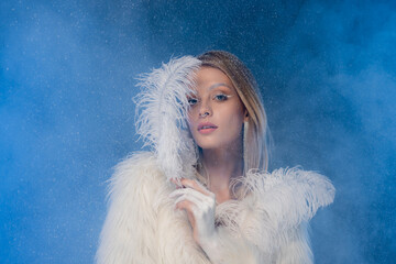 blonde woman with winter makeup in faux fur jacket holding white feather under falling snow on dark...