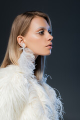 side view of blonde woman with winter makeup looking away and posing in faux fur jacket isolated on blue.