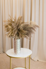 Soft home decor in gentle tones, a tall cylindrical vase with a bouquet of dried flowers against a background of beige curtains, on a round table. Interior.