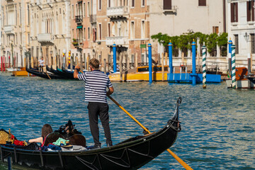 Fototapeta na wymiar A gondolier standing in a gondola controls the boat with an oar in his hands, a woman floats along the canal in a gondola along the coast of the street of the city of Venice, Venetian gondolier