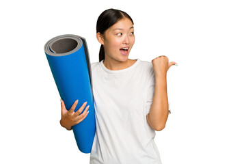 Young asian woman holding a mat isolated on green chroma background points with thumb finger away, laughing and carefree.