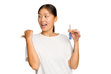 Young asian woman holding home keys isolated on green chroma background points with thumb finger away, laughing and carefree.