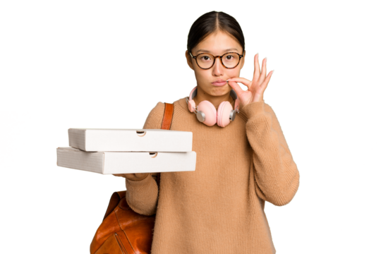 Young student Asian woman holding pizzas isolated on green chrome background with fingers on lips keeping a secret.