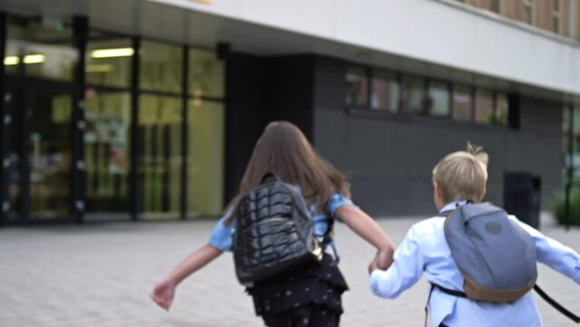 Two little students, a boy and a girl of 8-9 years old, say goodbye to someone and, holding hands, run to the school building.