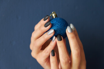 Obraz premium female hands with black and silver manicure on nails, hold a blue ball Christmas tree toy on a blue background