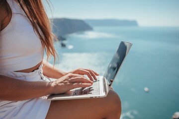 Digital nomad, woman in the hat, a business woman with a laptop sits on the rocks by the sea during sunset, makes a business transaction online from a distance. Freelance, remote work on vacation.