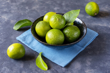 Group of fresh limes in dark blue bowl on blue napkin and on concrete background with leaves