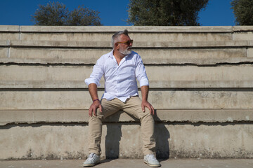 handsome mature man with beard and grey hair is sitting on the steps of an open-air auditorium. The man looks at different places. Senior concept, travel and tourism.