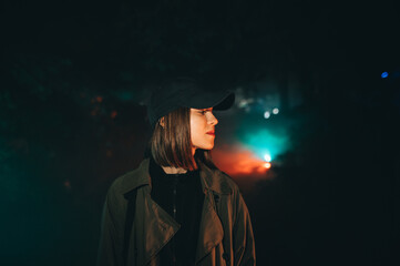 Evening portrait of a beautiful woman in casual clothes standing at night in a foggy park and looking away with a smile on her face.