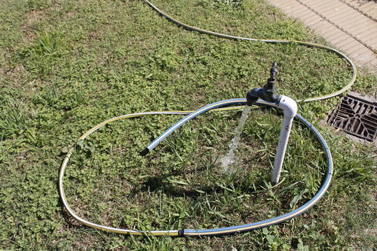 Manguera de riego de jard&iacute;n en el suelo con grifo de metal antiguo abierto que derrama agua, forma un dise&ntilde;o natural con fondo de hierba verde