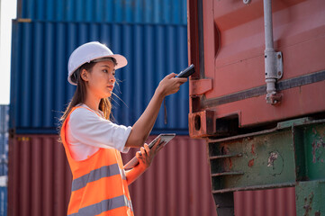 Smile Asian woman logistics worker wear safety helmet and protect suite working and checking product with taplets and scan code gun in shipping containerin commercial transport port