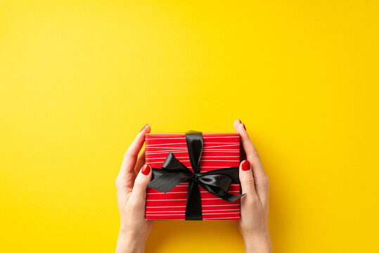 Black friday sales concept. First person top view photo of young woman's hands giving red gift box with black ribbon bow on isolated yellow background with empty space