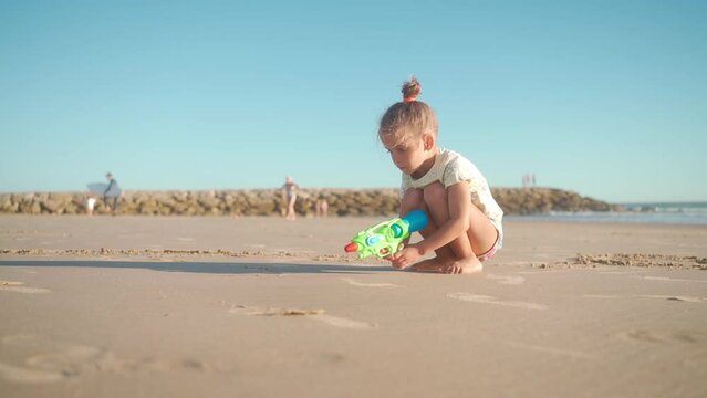 Child playing ocean beach summer day with water gun. Little girl caucasian have fun run on sand seashore with squirt gun. Adorable female 5 years old enjoy playing on beach Handheld. 