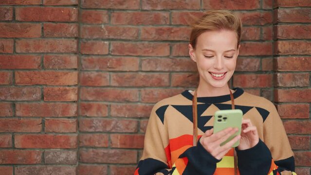 Handsome blonde woman typing on phone near brick wall outdoors