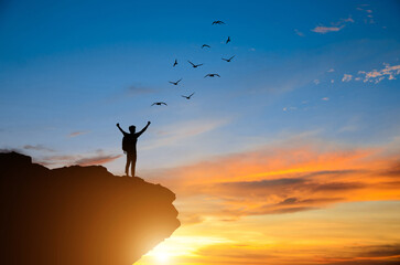 Silhouette of young man standing alone on top of mountain and raise both arms praying and free bird...