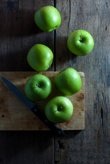 Green apples with a kitchen knife and wooden cutting board on a rustic wooden floor, top view.