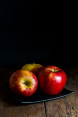 Red apples are placed on a black square ceramic plate on a rustic wooden floor.