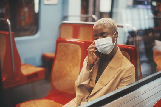 A Portrait With A Shallow Depth Of Field Of A Young Black Lady With Short Hair Painted In White Color, With A Mask On Her Face, Sitting Alone On A Red Seat Of A Subway Train And Talking On The Phone