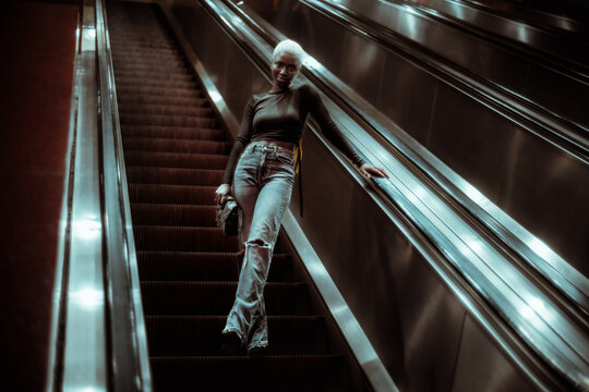 A Low-key Shot Of A Young Cute Black Female Standing Alone On The Moving Staircase And Descending To The Metro Station; A Youthful African Woman Is Getting Down To A Subway Platform Via Escalator
