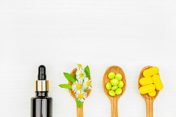 Pills in wooden spoons, natural oil bottle and medical plant on white background