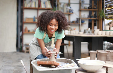 Close-up of concentrated beautiful craftswoman in apron sitting at pottery wheel and using craft tool while shaping wet clay vessel