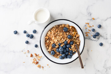 Bowl of granola with blueberries and jar of dairy free milk on white marble table background, top...