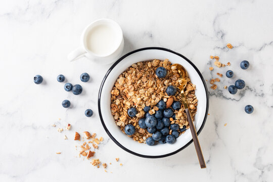Bowl of granola with blueberries and jar of dairy free milk on white marble table background, top view