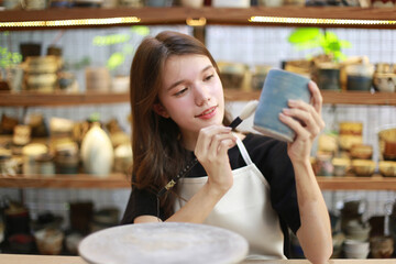 Close-up of concentrated beautiful craftswoman in apron sitting at pottery wheel and using craft tool while shaping wet clay vessel