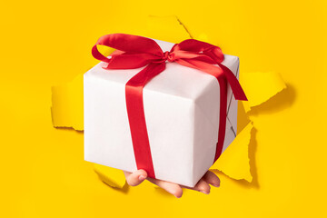 Hand giving gift box through torn yellow paper background, closeup. Special offer or sale concept