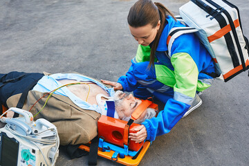 Female paramedic fixing head of male victim with neck injury lying on ambulance stretcher. First aid with cervical collar and defibrillator - obrazy, fototapety, plakaty